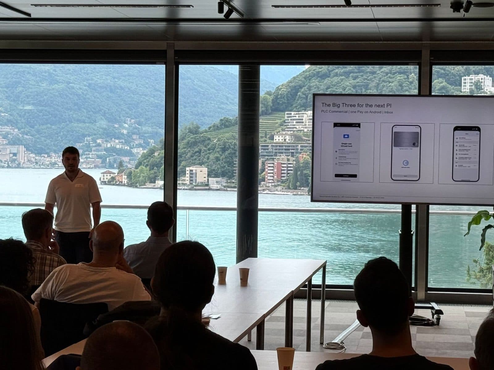 Florian Raschle presenting to an audience, three phone screens about virtual cards on the display behind him, a lake in the background